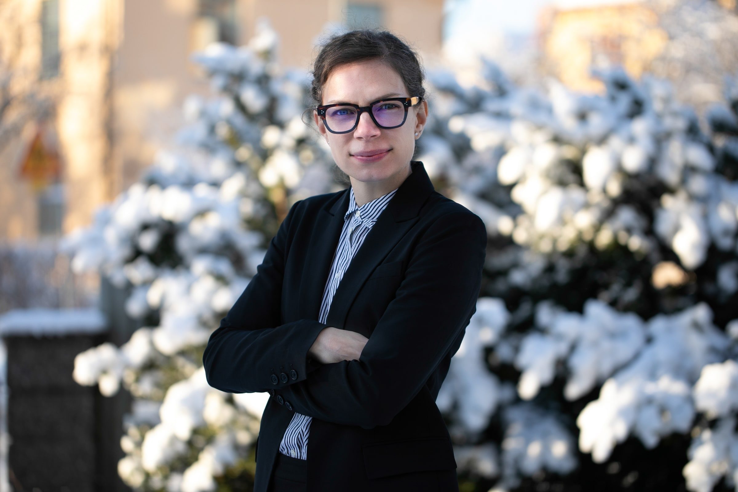Team member in glasses and a dark blazer standing with folded arms outdoors.
