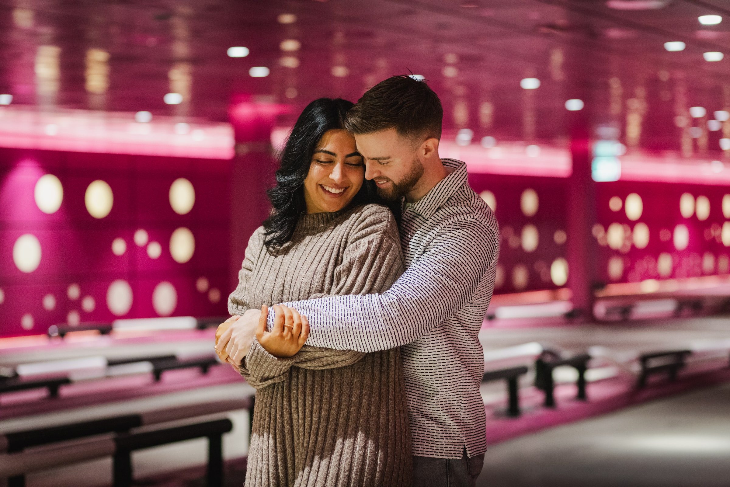 Couple embracing beneath pink lights in a games venue.