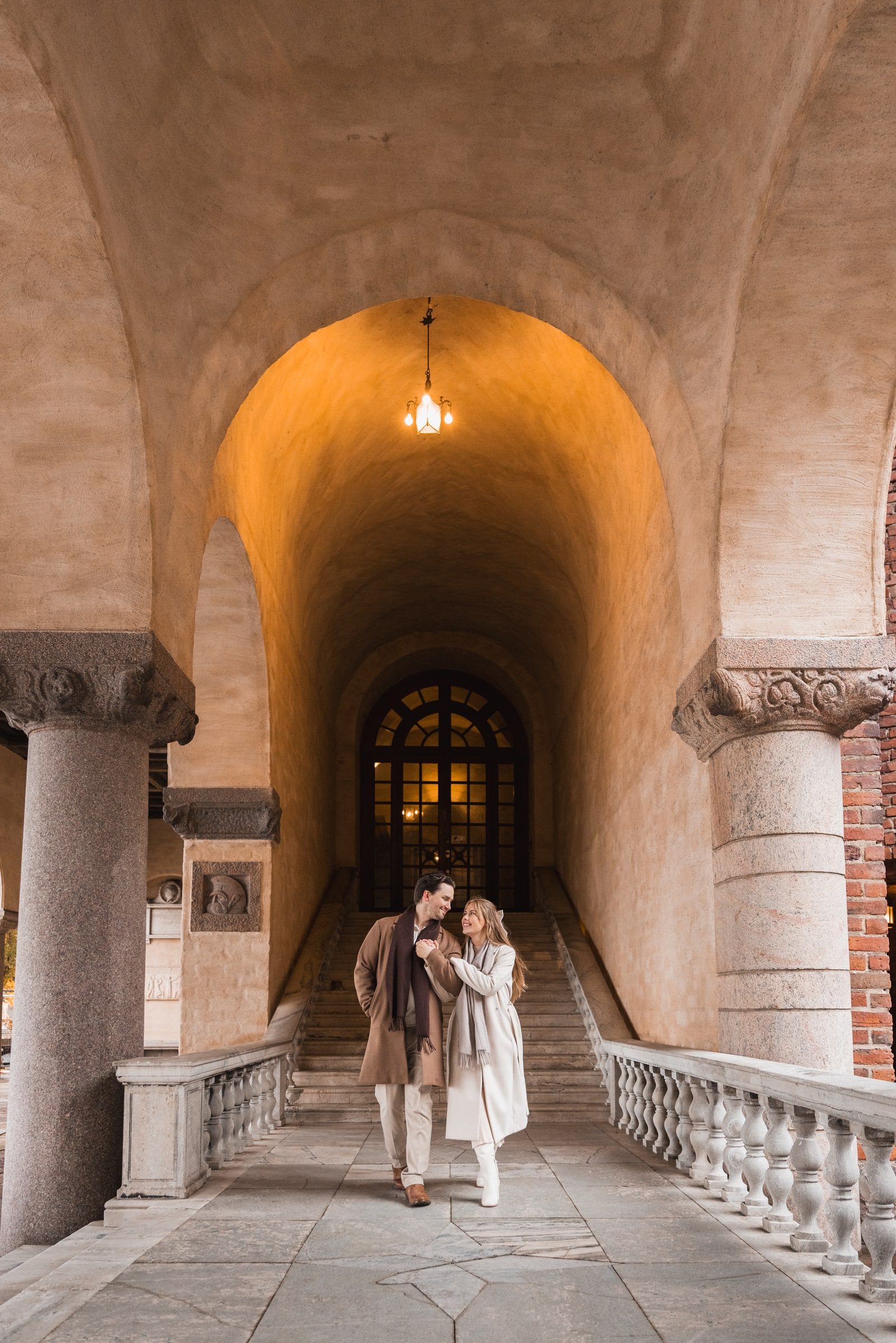 Couple standing beneath tall stone arches in a quiet architectural portrait.