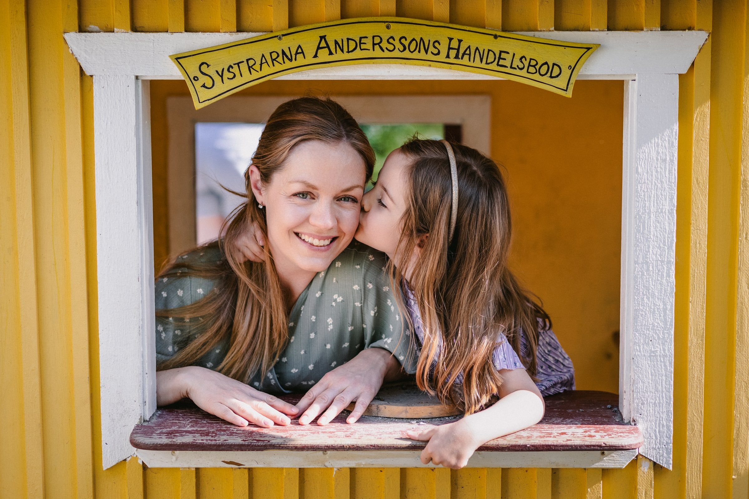 Mother and daughter sharing a playful moment at a small yellow playhouse window.