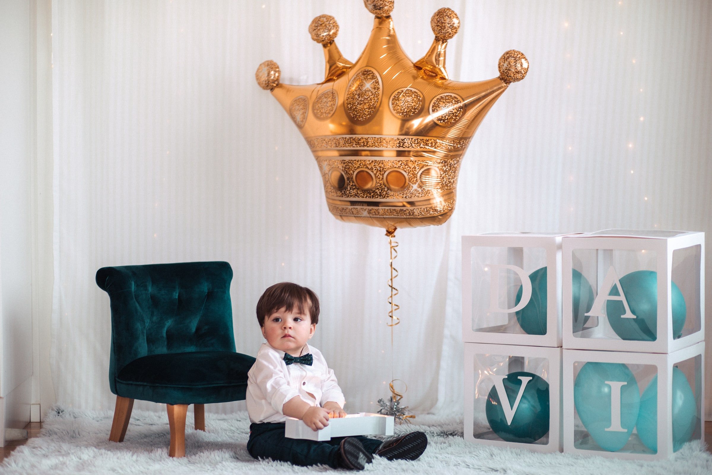 Baby seated beneath a gold crown balloon beside first-birthday letter blocks.