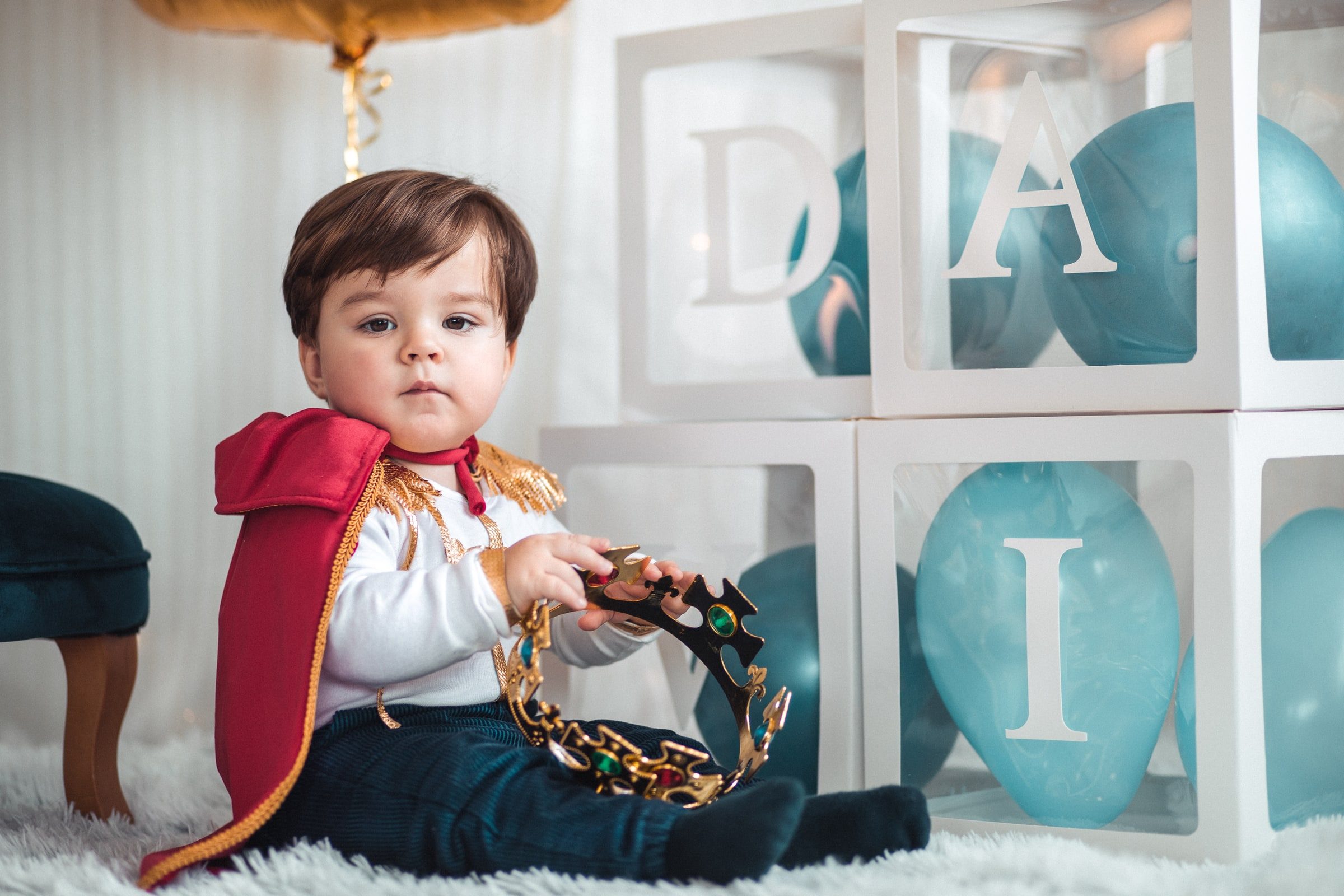 Baby in a red cape holding a tambourine beside the birthday decorations.